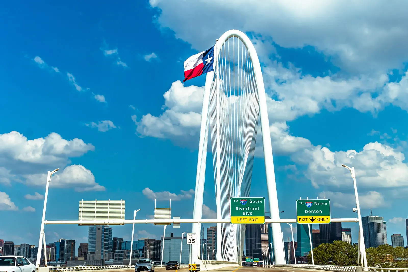 The Texas flag flying over the Margaret Hunt Hill Bridge with the Dallas skyline behind it
