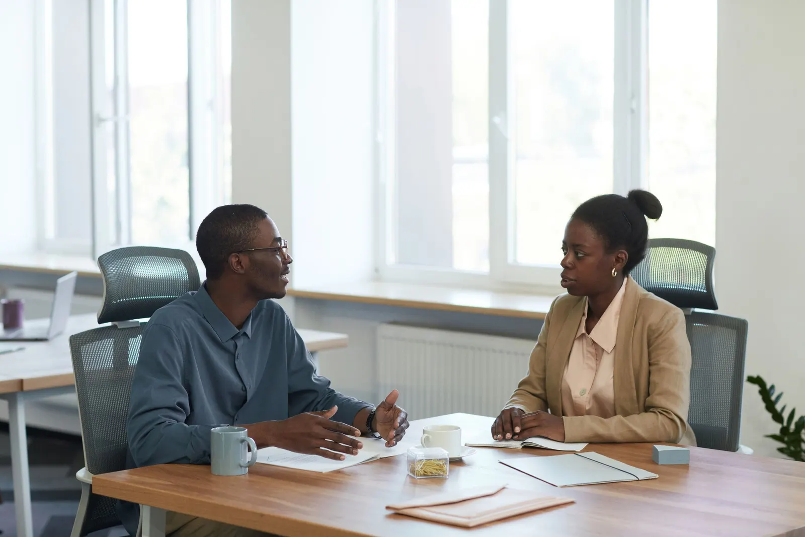 Two people talk across a desk in a bright office