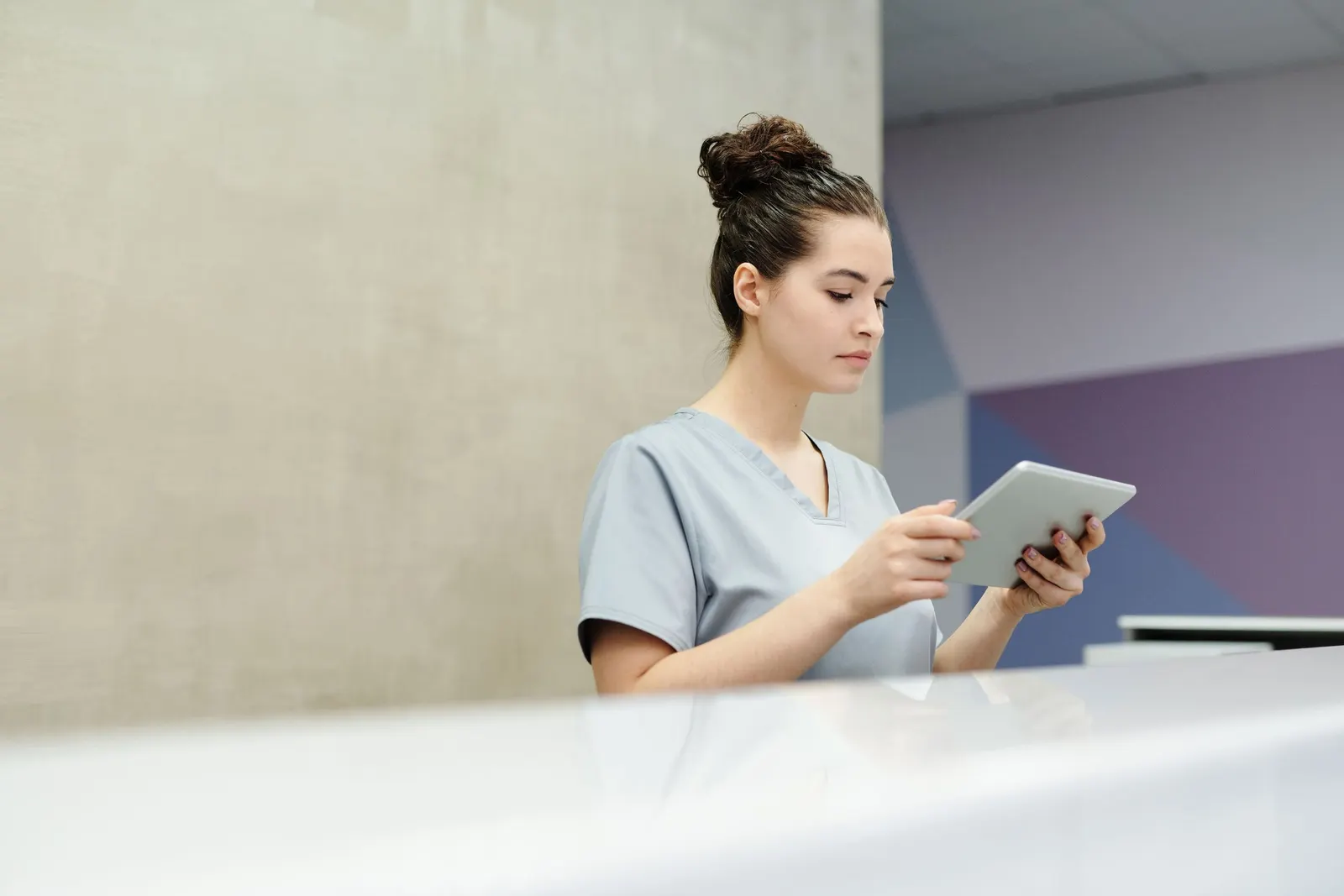 A receptionist in scrubs checks a tablet at a clinic front desk