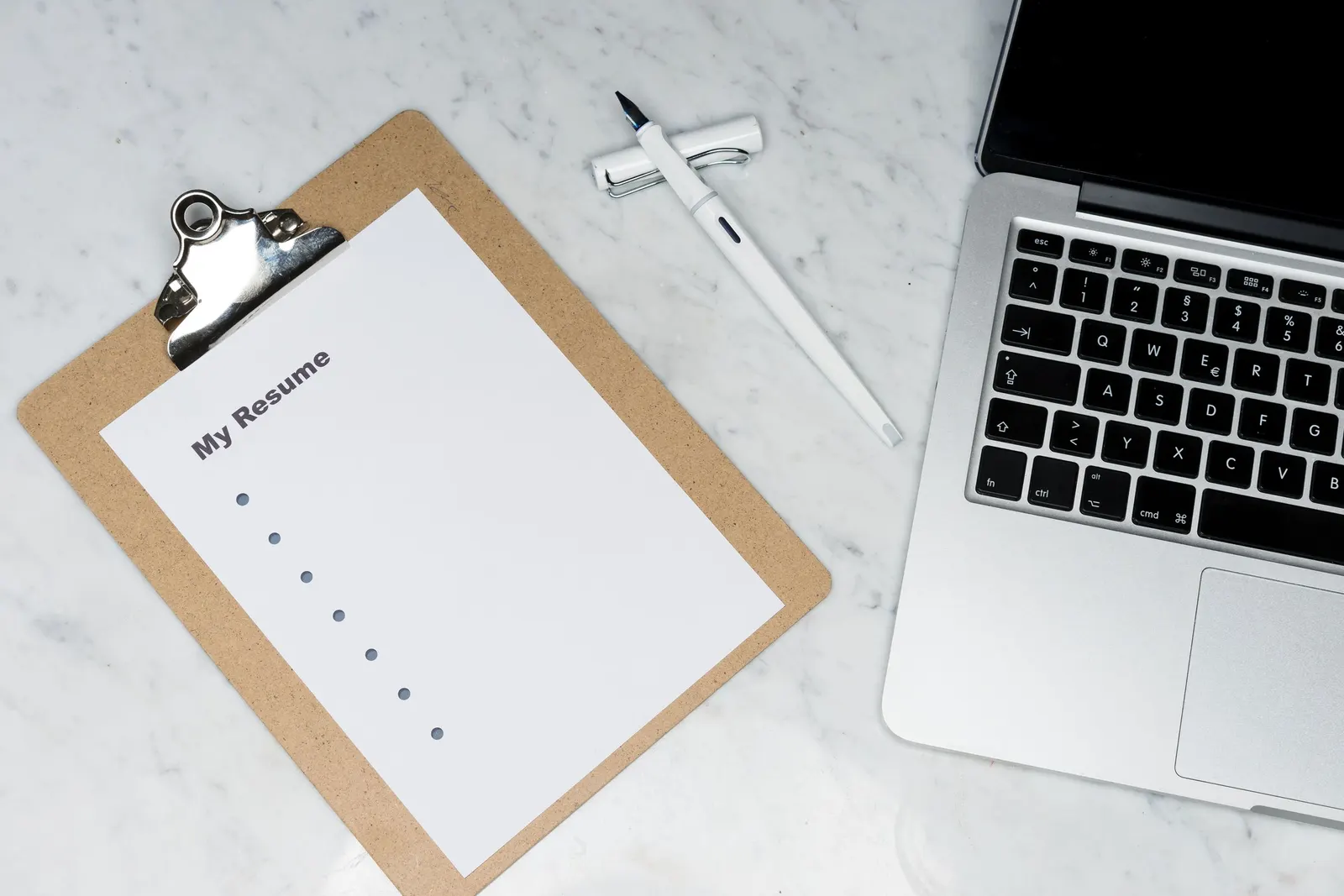 A resume on a clipboard beside a pen and a laptop on a desk