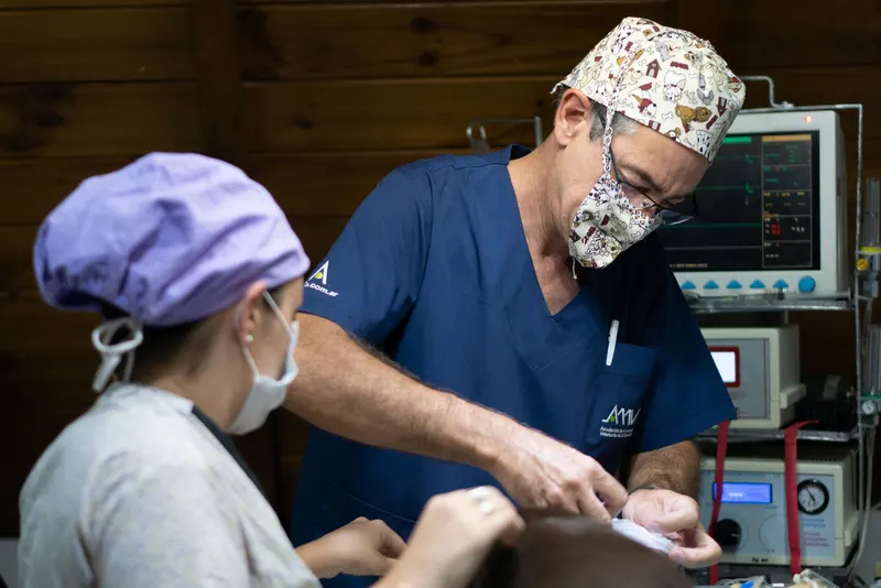 Two veterinary technicians in scrubs and masks work at a treatment table beside a patient monitor